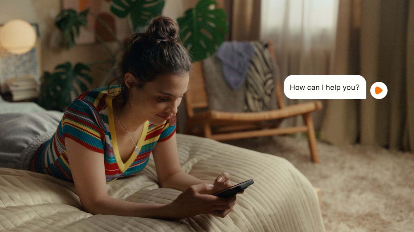 A figure lies on a bed with their phone in hand. A pop-up white circle on the right of the image has a speech bubble to its left, which reads, "How can I help you?".