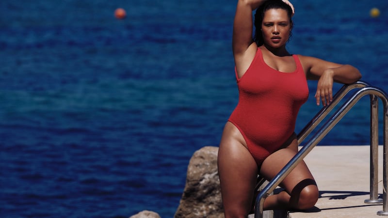 A model poses with her arm above her head in a red crinkle Hunza G swimsuit against a sea backdrop.